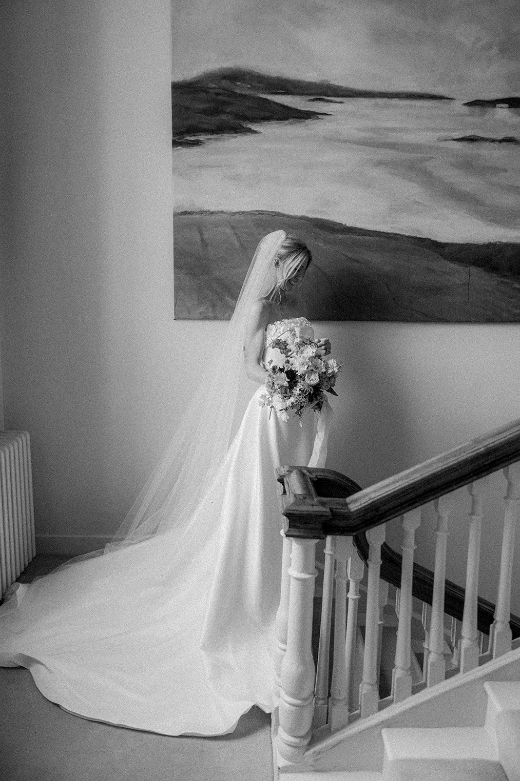 A bride in a wedding dress and veil holding a bouquet of flowers on a staircase landing indoors, with an abstract landscape painting on the wall behind her.