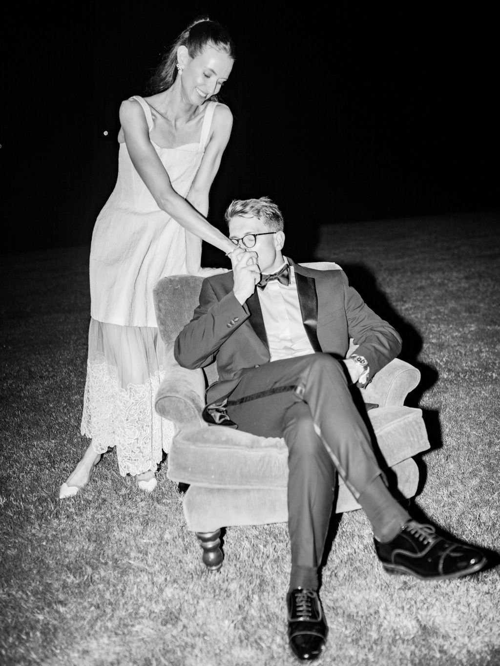 A woman in a dress playfully holds a man's nose, who is seated in an armchair, wearing a tuxedo, glasses, and a bow tie, outdoors at night.