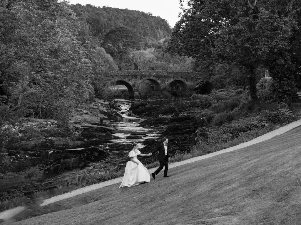 A bride and groom walking on a grassy slope next to a river with trees and an arched stone bridge in the background, in black and white.