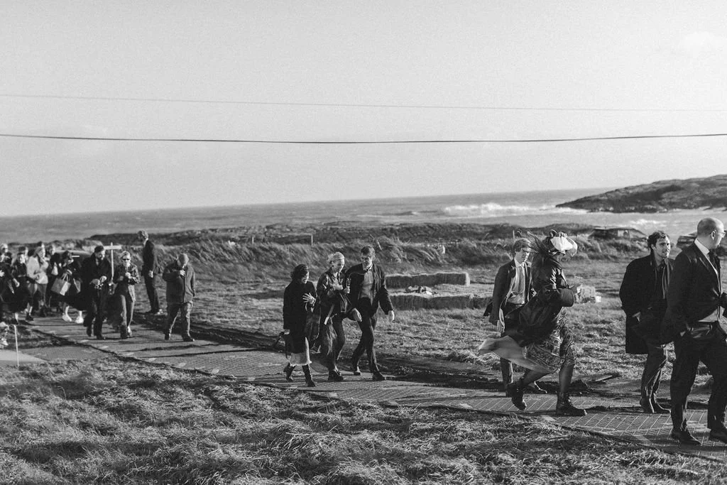 A group of people walking along a coastal path near the ocean, some wearing masks and holding papers.