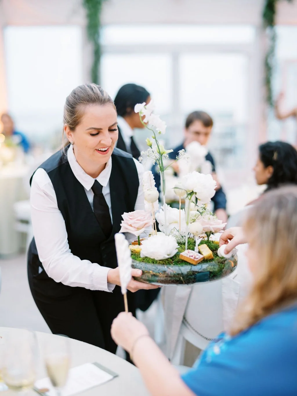 A server carrying a floral centerpiece with white and pale pink flowers at a reception event.