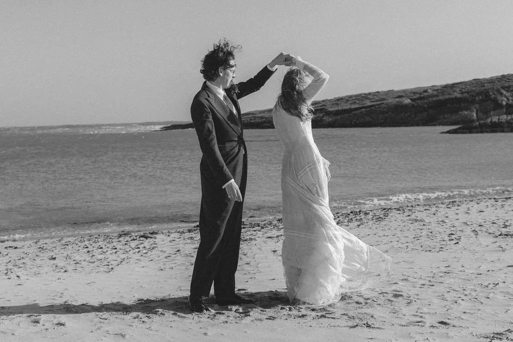 A couple dancing on the beach, with the man in a suit and the woman in a flowing dress, holding hands and spinning.