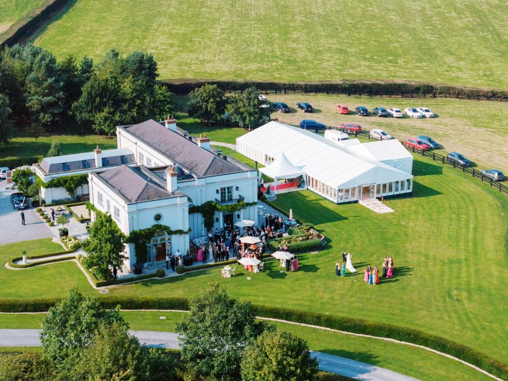 A large estate with a white mansion and a big white event tent on a lush green lawn, with guests gathered outside and several parked cars.