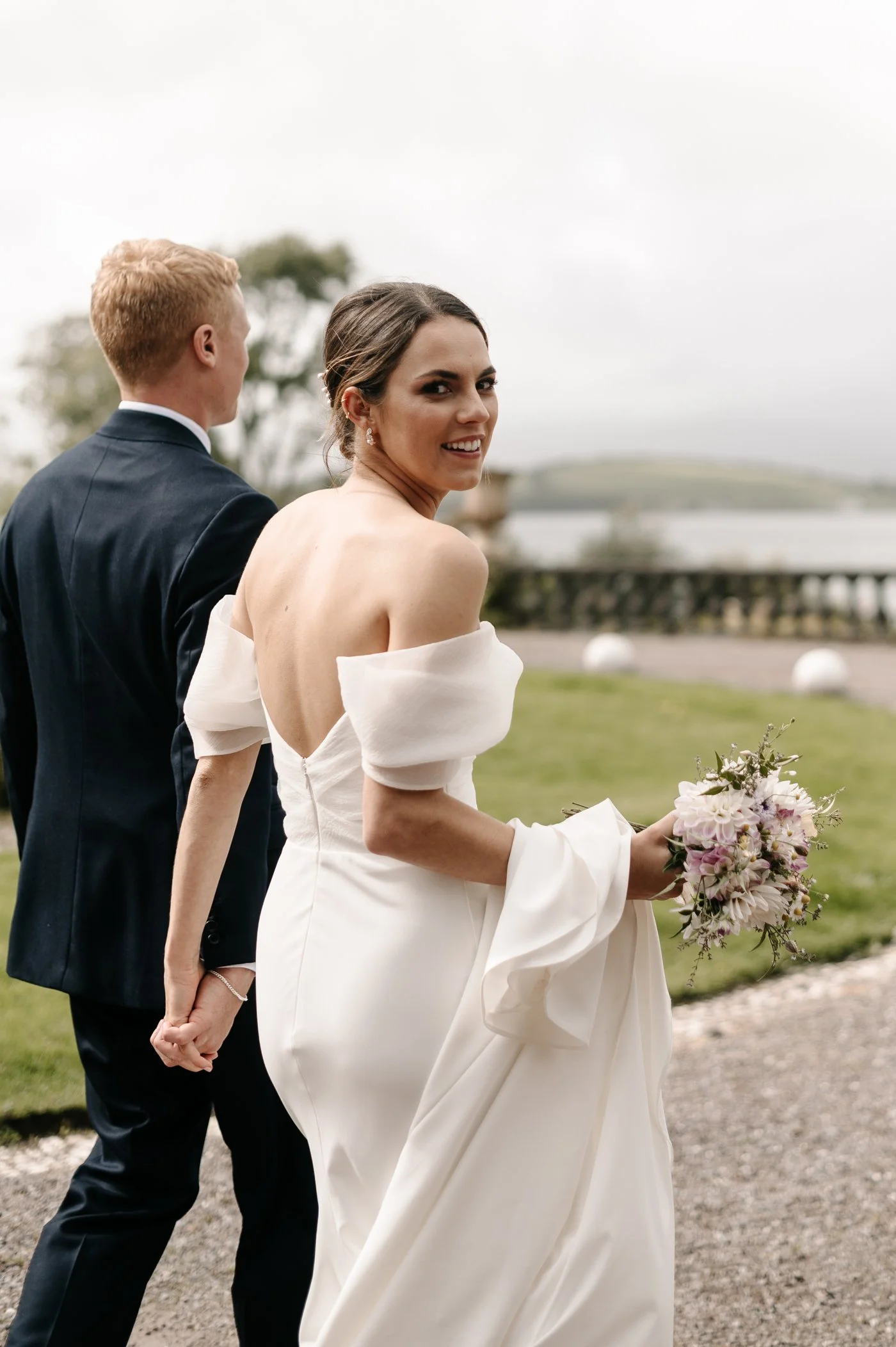 A bride in a white wedding dress holding a bouquet of flowers look back at the camera, while a groom stands beside her, facing away. They are outdoors near water with cloudy skies.