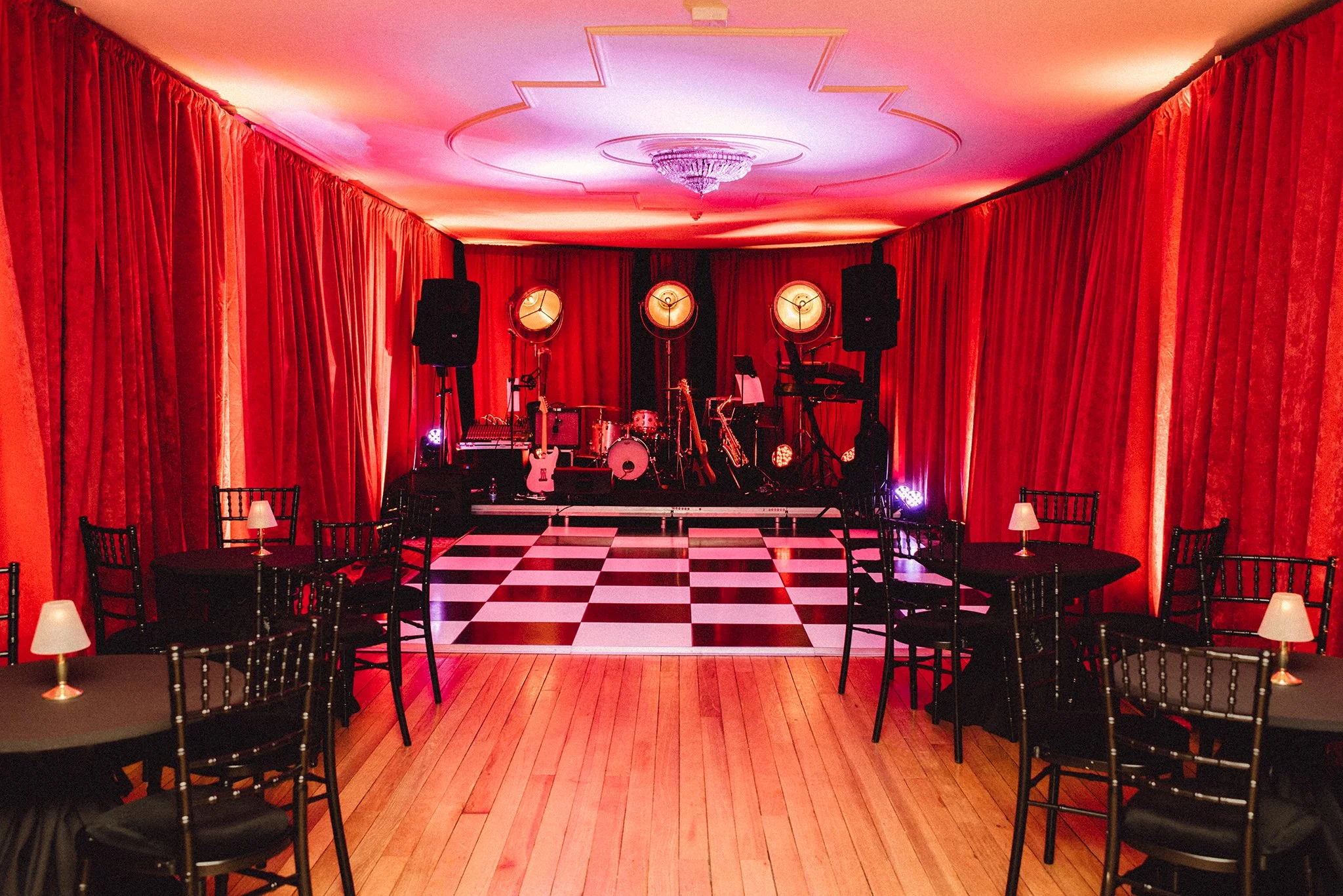 An empty performance stage with musical instruments, including drums, guitars, and keyboards, in a dimly lit room with red curtains and chairs, small tables, and lamps.