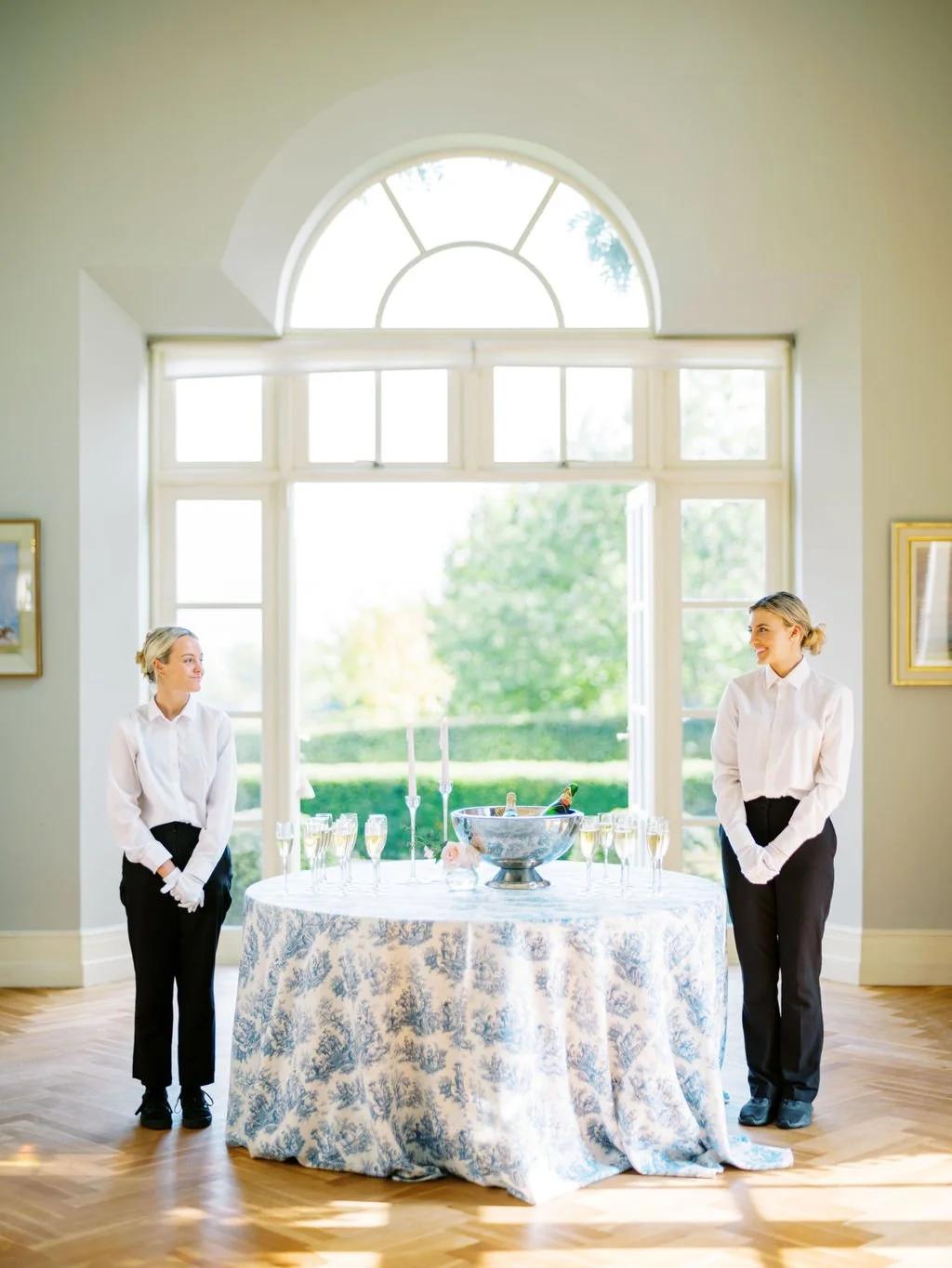 Two women in white shirts and black pants standing on either side of a round table with a blue and white floral tablecloth, champagne glasses, tall candles, and a champagne bucket, in front of large windows with greenery outside.
