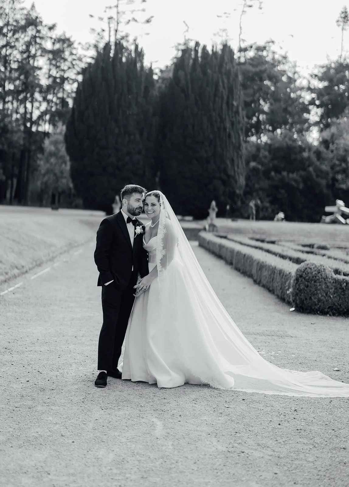 Black-and-white photo of a bride and groom standing on a garden path, smiling. The groom wears a black tuxedo, and the bride wears a white wedding gown with a long veil. Large trees and trimmed bushes are in the background.