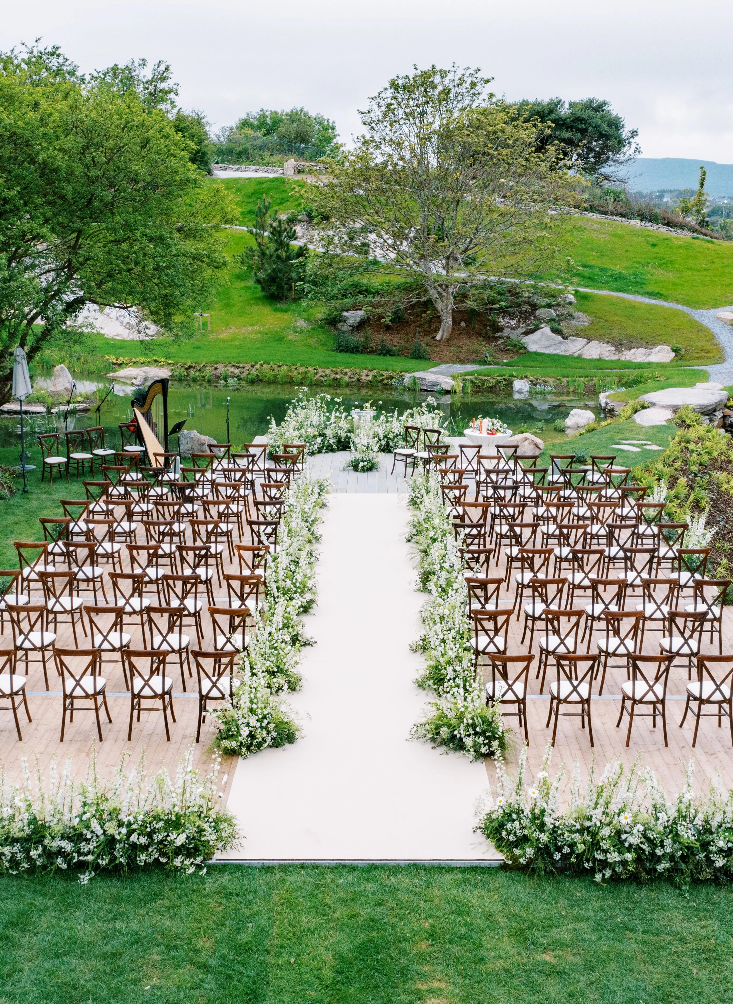 Outdoor wedding ceremony setup with rows of wooden chairs on a white aisle runner, floral arrangements along the aisle, near a pond with greenery and trees in the background.