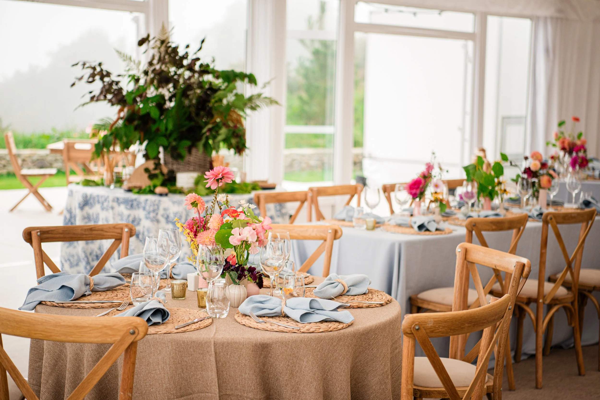 Elegant indoor dining setup with round tables, each decorated with floral centerpieces, blue napkins, glassware, and plates, surrounded by wooden chairs, with large windows and outdoor greenery in the background.
