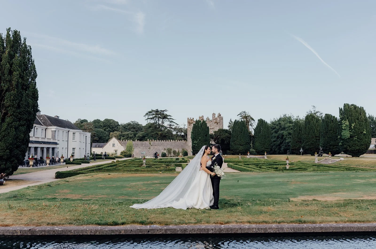 Bride and groom by the water with the castle ruins behind them at Castlemartyr Resort, Cork