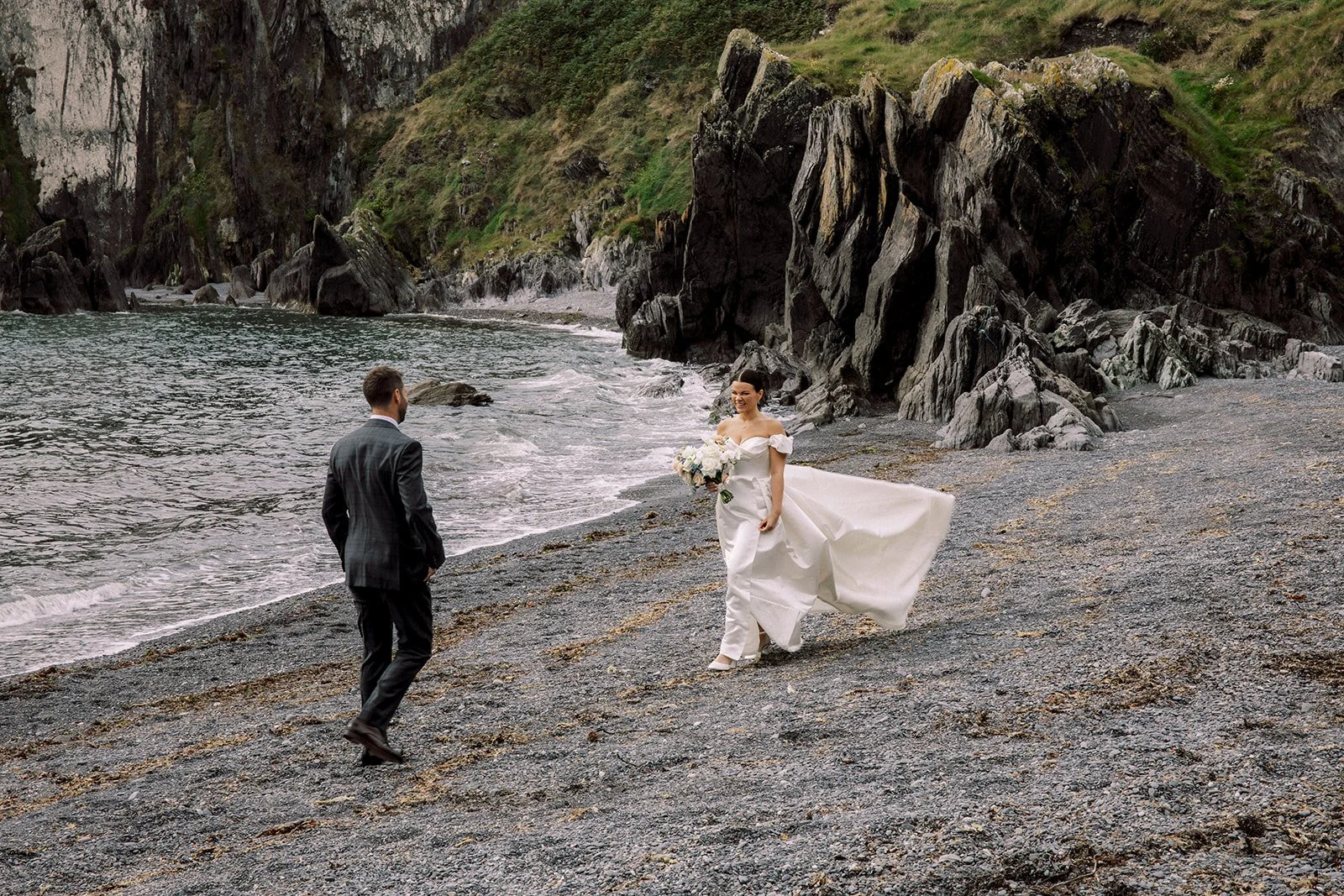 A bride in a white wedding dress holding a bouquet, smiling and walking on a rocky beach, while a groom, dressed in a dark suit, walks towards her. Large rock formations and green hills are in the background.