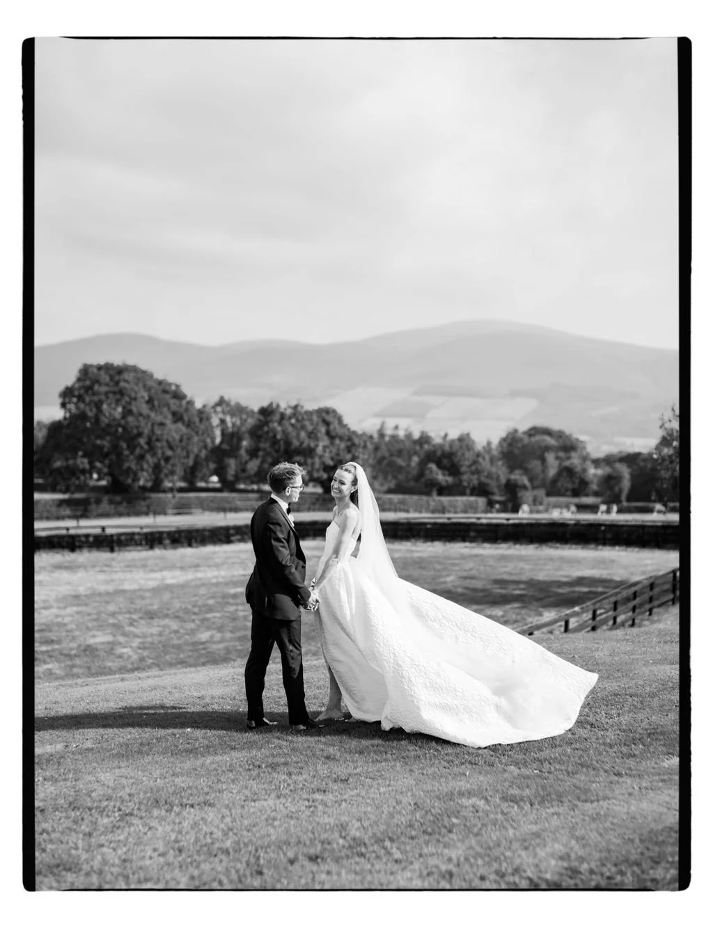 Black and white photo of a bride and groom holding hands and smiling outdoors on a grassy field with trees and mountains in the background.