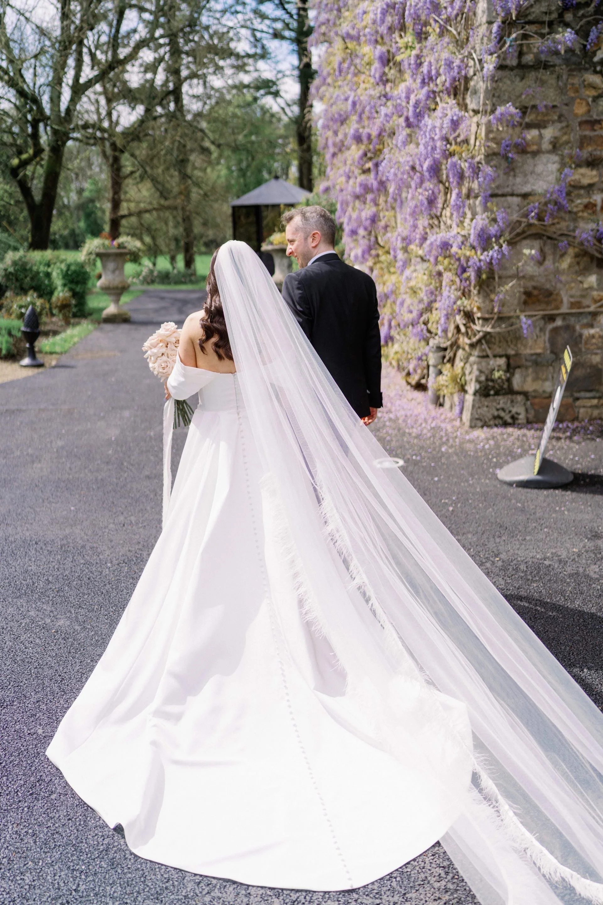 Bride in a white wedding gown and veil holding a bouquet, walking with groom in a black suit along a garden pathway with purple flowering vines on a stone wall.