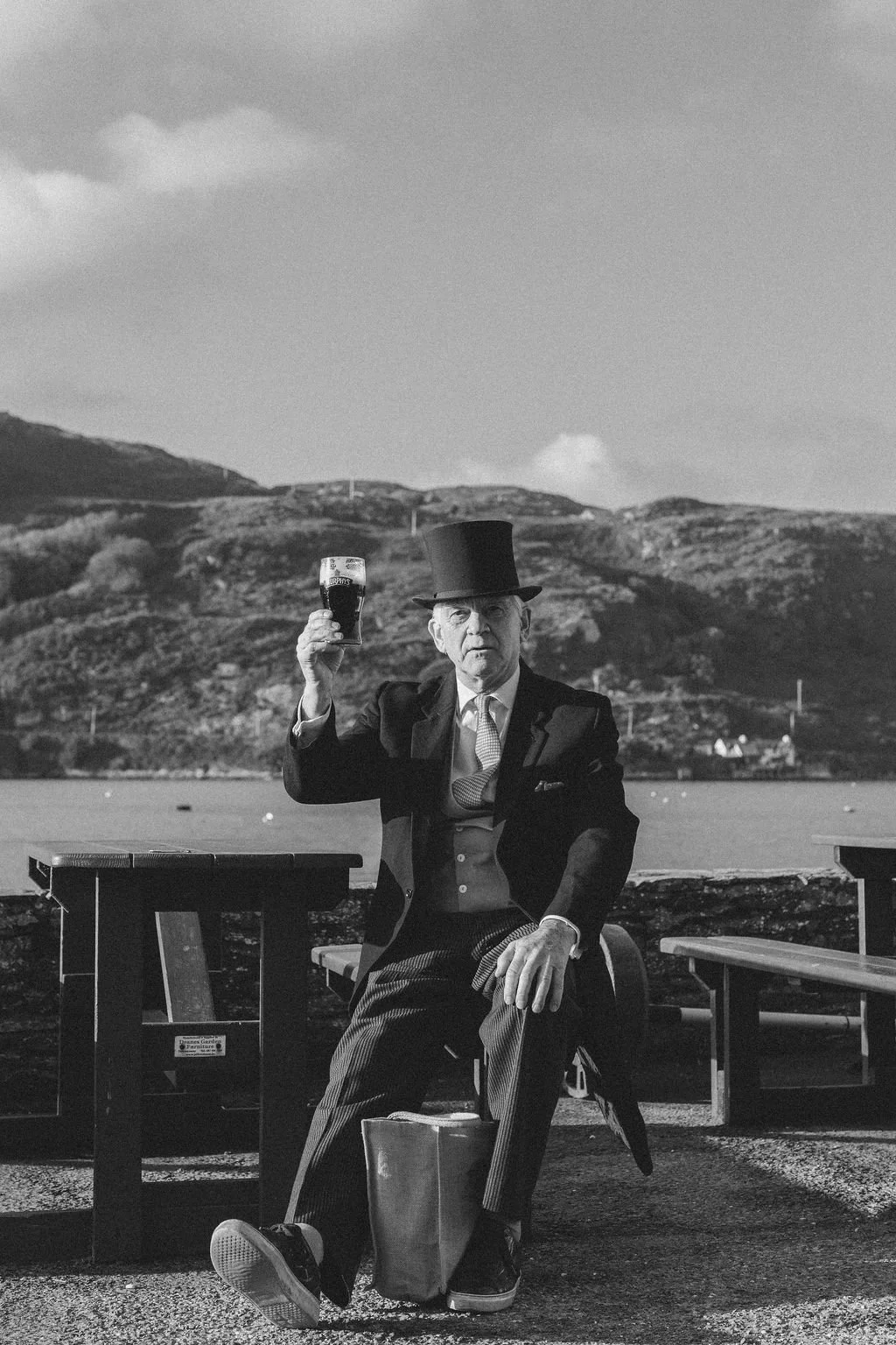 Black and white photo of an elderly man wearing a top hat, sitting outdoors by a lake with hills in the background, holding a glass of dark beer and dressed in a suit.