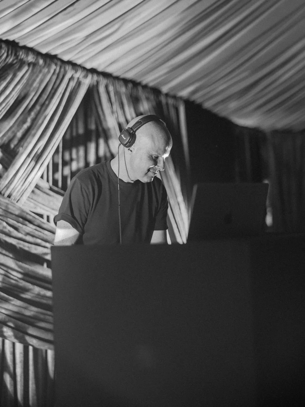 A man wearing headphones standing at a DJ booth with a laptop, in a space with bamboo walls and curtains.
