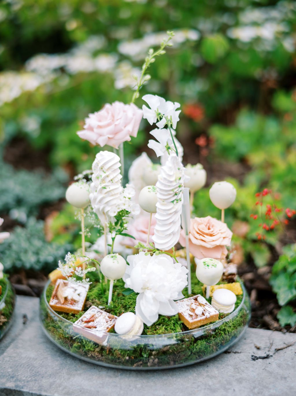 A dessert display with white and cream-colored sweets, pink roses, and white flowers set in a round glass dish on a gray surface with a blurred green garden background.
