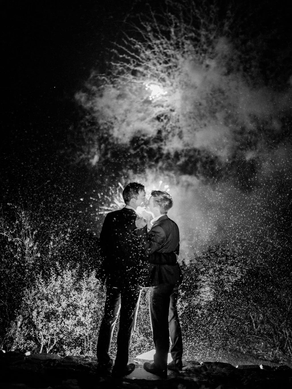 Two men in suits embracing at night with fireworks in the sky behind them.