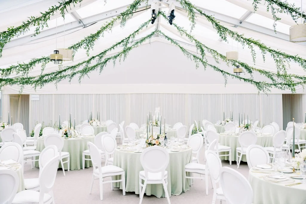 Elegant event tent with white and green color scheme, decorated with hanging greenery, round tables covered with light green tablecloths, white chairs, and floral centerpieces.