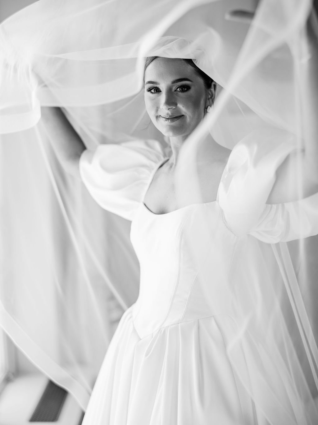 A woman in a wedding dress with a veil, smiling, and looking at the camera, surrounded by flowing fabric in a black-and-white photograph.