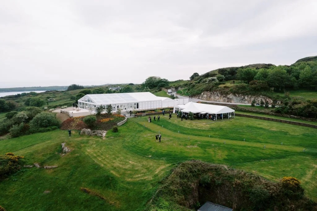 A large white tent and a greenhouse-like structure set up on a green, grassy area with hills and trees in the background, likely for an outdoor event or gathering.