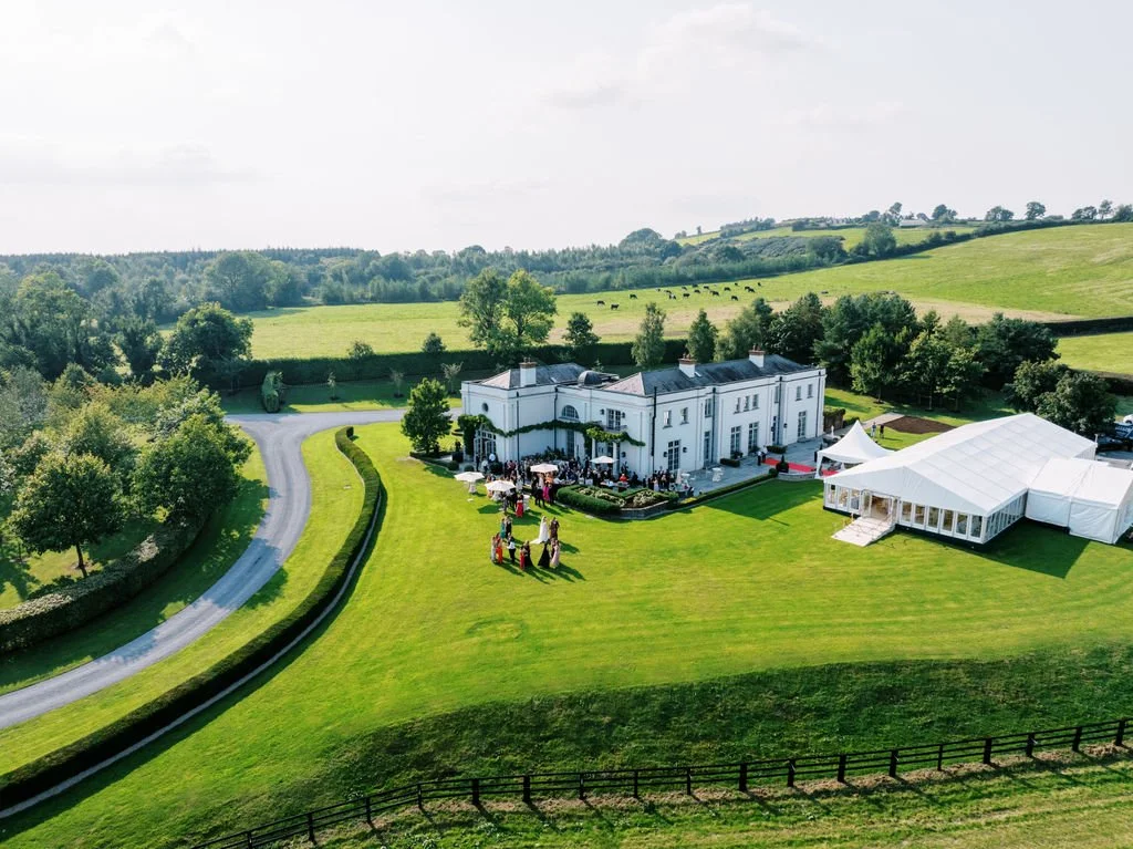 An aerial view of a large white estate with outdoor event seating, a white tent, and lush green lawns surrounded by rolling hills and trees.