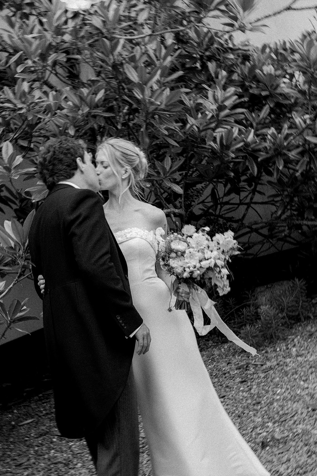 A black and white photo of a bride and groom sharing a kiss outdoors. The bride is holding a large bouquet of flowers, and they are standing in front of dense foliage.