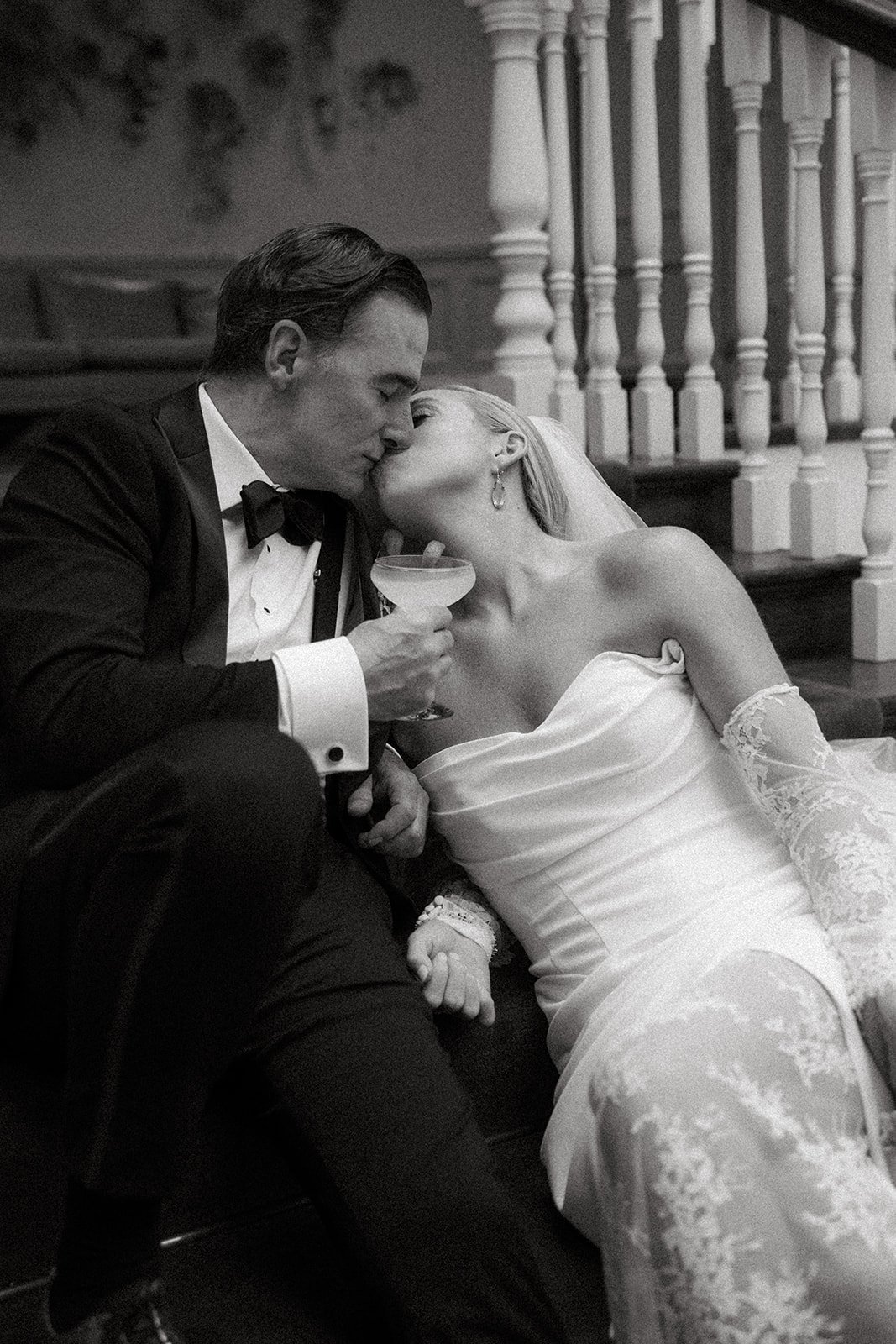 A black and white photo of a bride and groom kissing on a staircase, with the bride holding a cocktail glass.