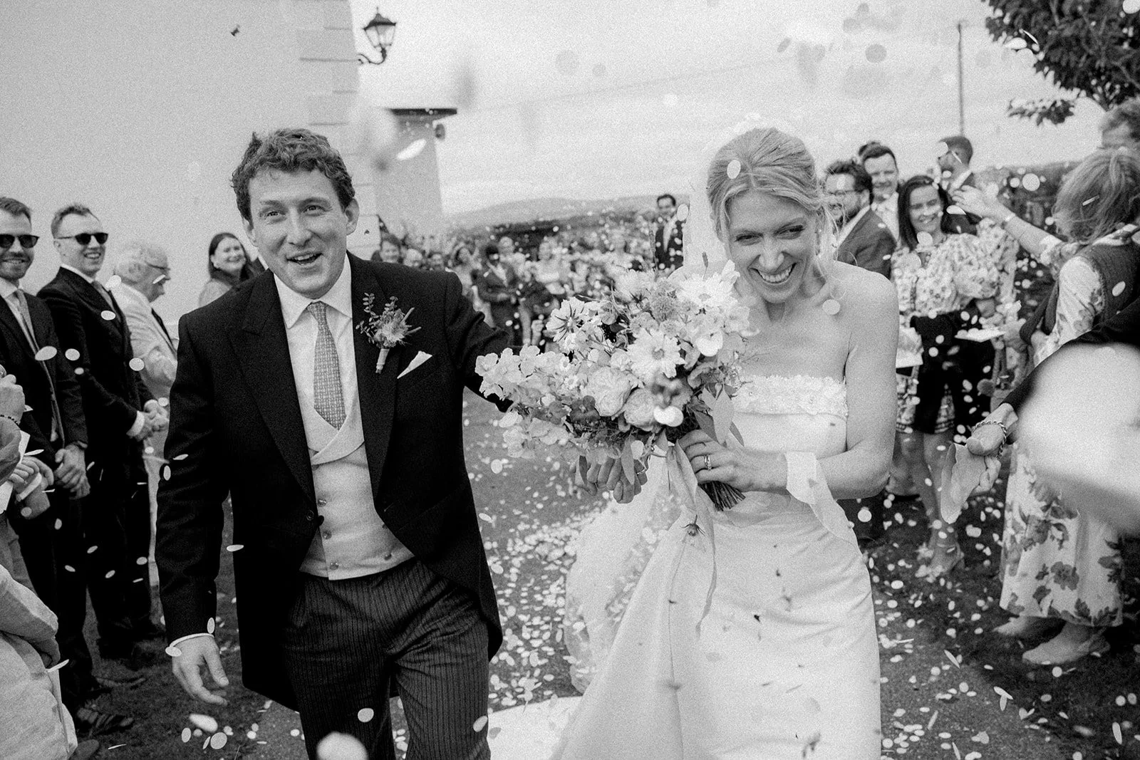 Black and white photo of a bride and groom walking outside with confetti, surrounded by happy guests.