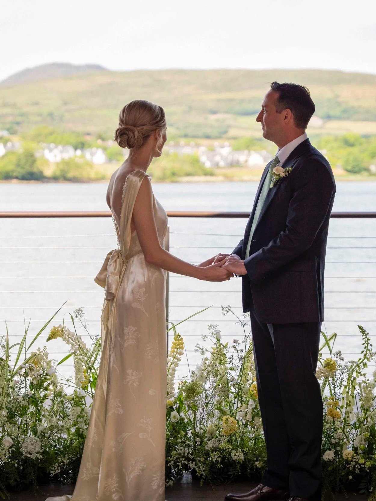A wedding ceremony with a bride and groom holding hands and facing each other, standing in front of a window overlooking a body of water and green hills, with floral decorations at their feet.