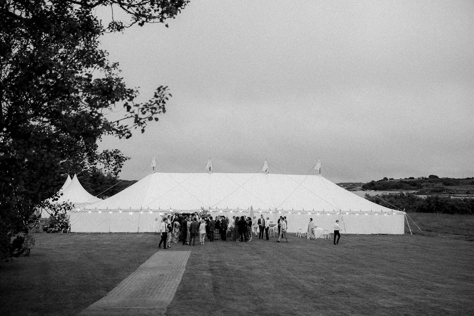 Black and white photo of a large event tent with a group of people gathered outside, situated on a grassy field with trees on the left and open landscape in the distance.