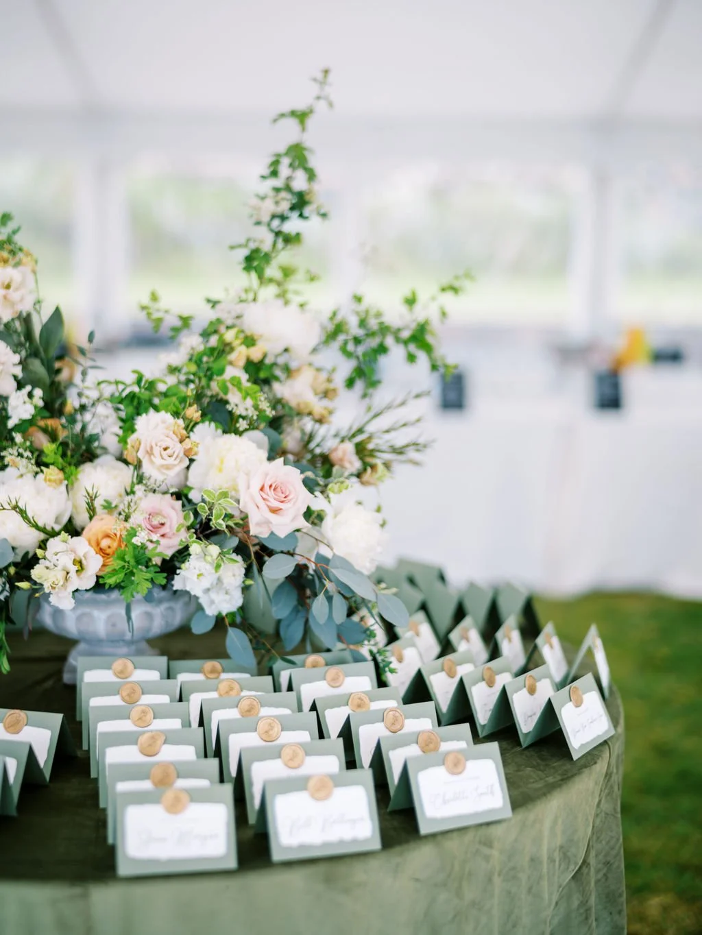 Table with small place cards sealed with wax stamps and a large floral arrangement of white, pink, and orange roses, along with greenery, inside a bright tent or pavilion.