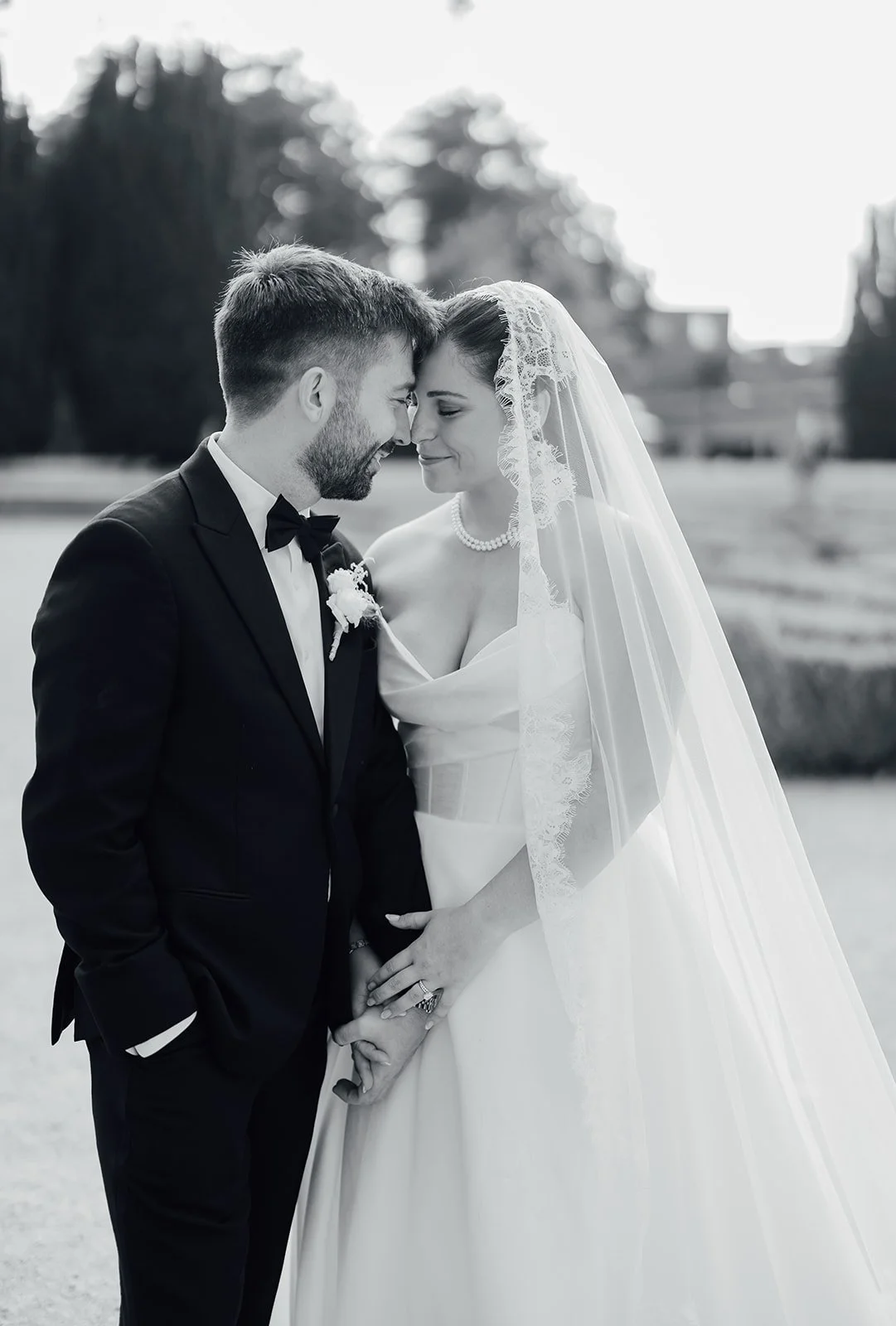 Black and white photo of a bride and groom on their wedding day, touching foreheads and holding hands, outdoors with trees in the background at Castlemartyr Resort