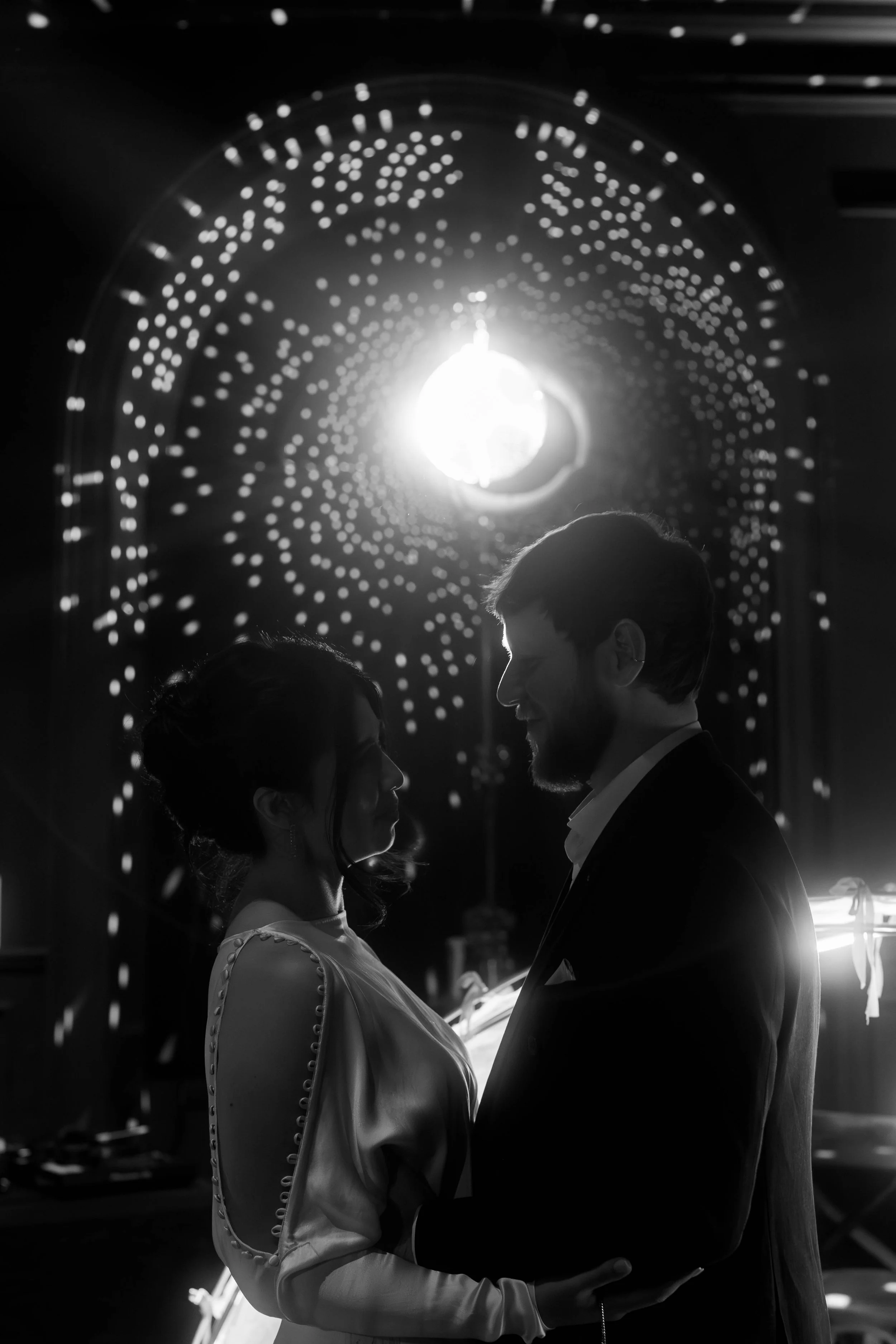 Silhouette of a couple dancing in front of a bright light with circular pattern in the background, in black and white.