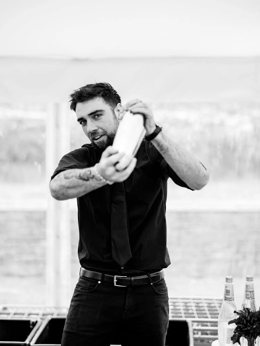 A man with dark hair and tattoos on his arm taking a selfie with a smartphone in an outdoor setting, with bottles on a table in the background.