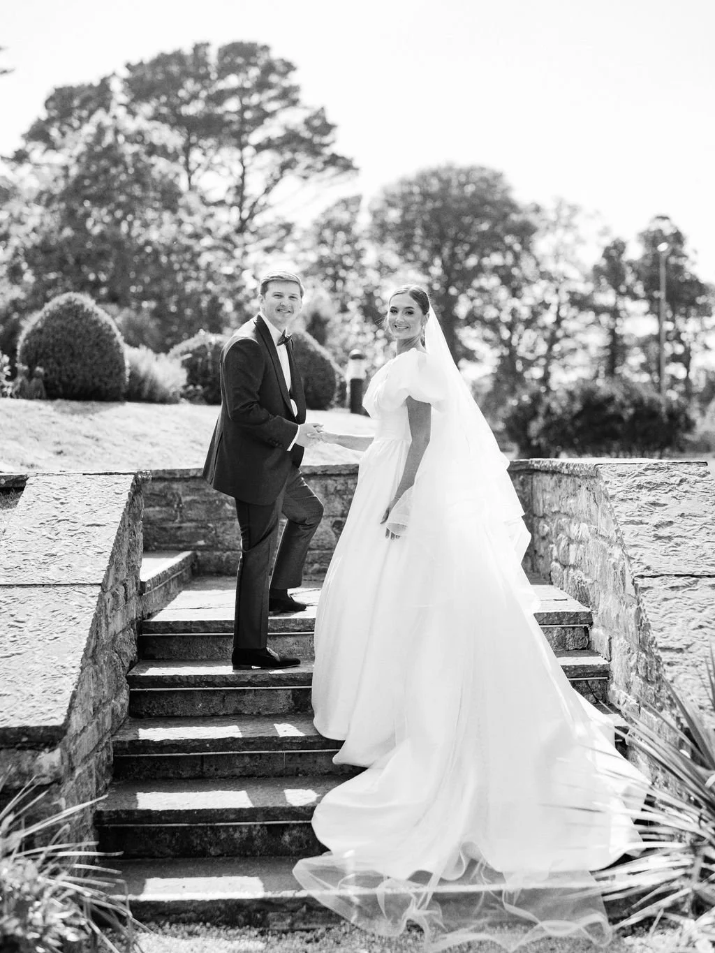 Black and white photo of a bride and groom holding hands on outdoor stone stairs with trees in background.