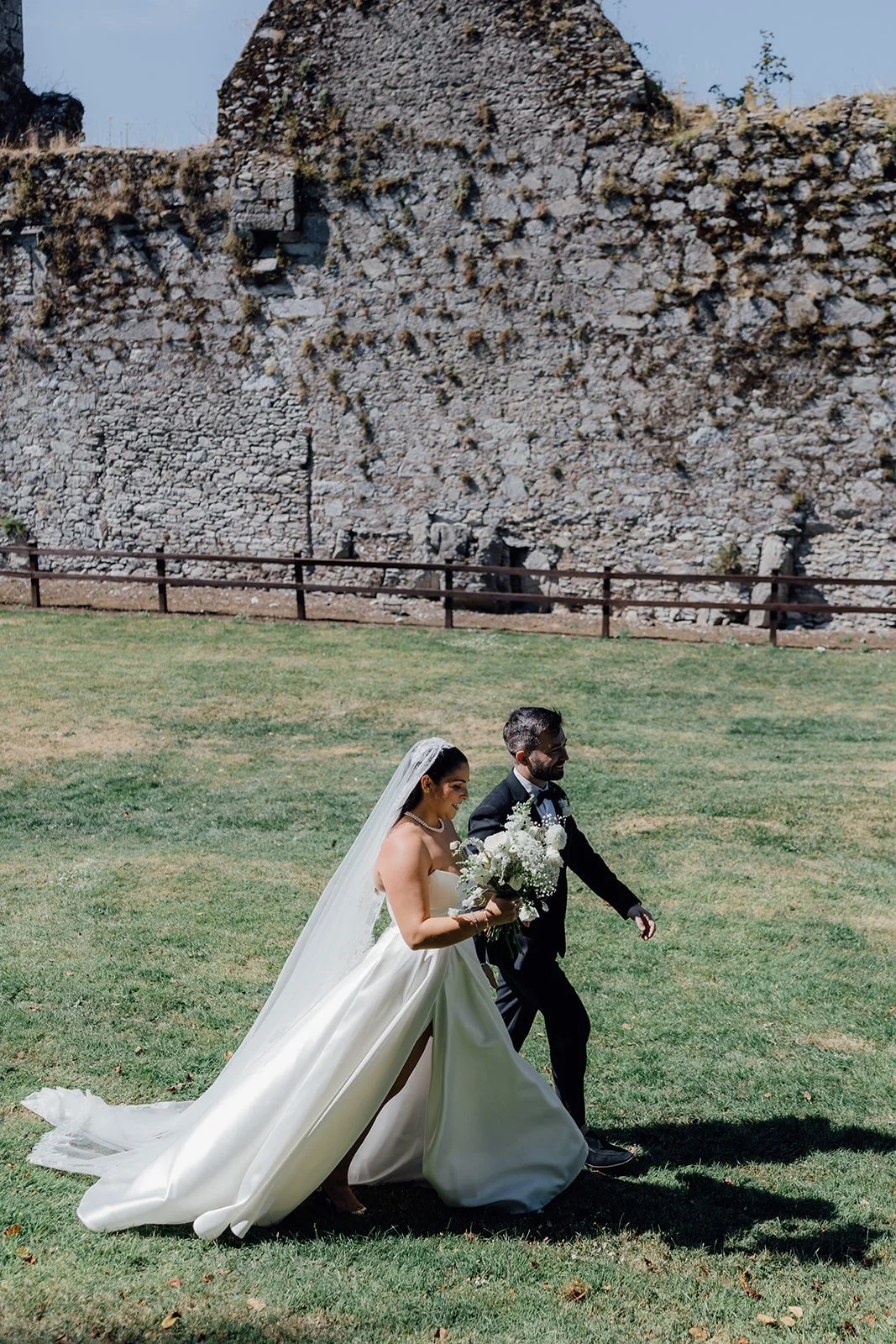 A bride and groom walking together outdoors on a sunny day, with the bride holding a bouquet of white flowers and wearing a white wedding gown with a veil, and the groom in a black suit with a white shirt and black bow tie, in front of a large stone wall.