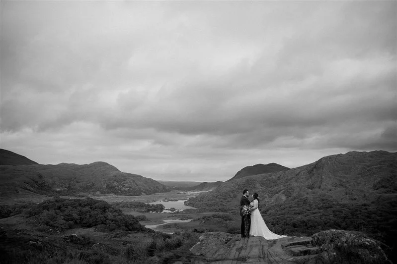 A black and white photo of a bride and groom standing close together on a rock overlooking a valley with mountains and a river.