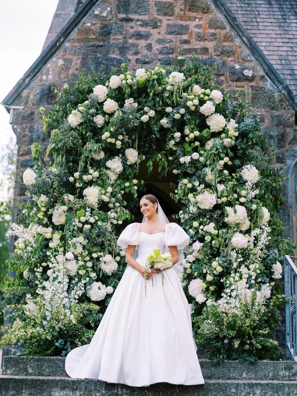 Bride standing outside a stone church entrance decorated with white flowers, wearing a white wedding dress and holding a bouquet of white flowers.