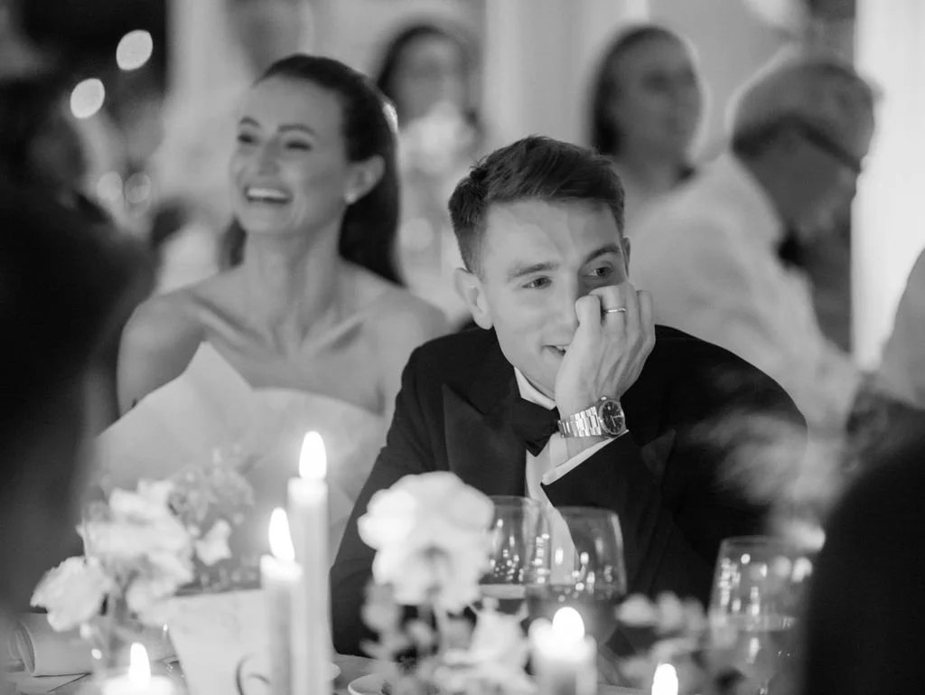 People sitting at a table during a formal celebration, with a woman laughing and a man wearing a tuxedo covering his mouth, all in a black-and-white photo.