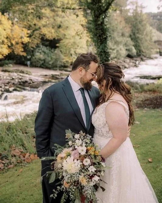 A newlywed couple stands closely together, with foreheads touching, outdoors near a river, surrounded by trees with fall foliage. The groom wears a dark suit and tie, and the bride wears a white lace wedding dress and holds a large bouquet of flowers.