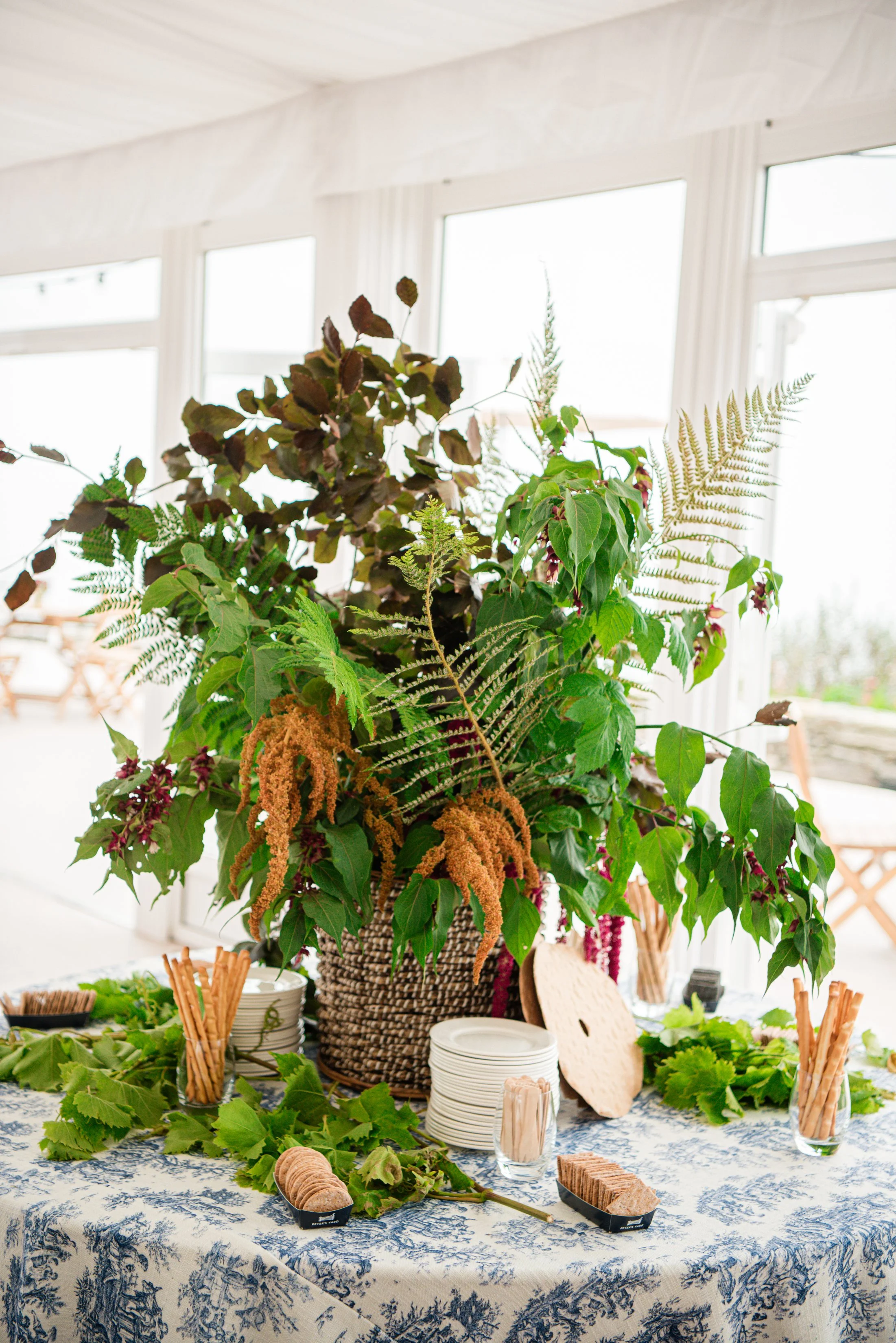 A large floral arrangement with various green and brown leaves and ferns, placed in a wicker basket on a table with a blue and white patterned tablecloth. The table has plates, crackers, and leafy garnishes, in front of a bright window.