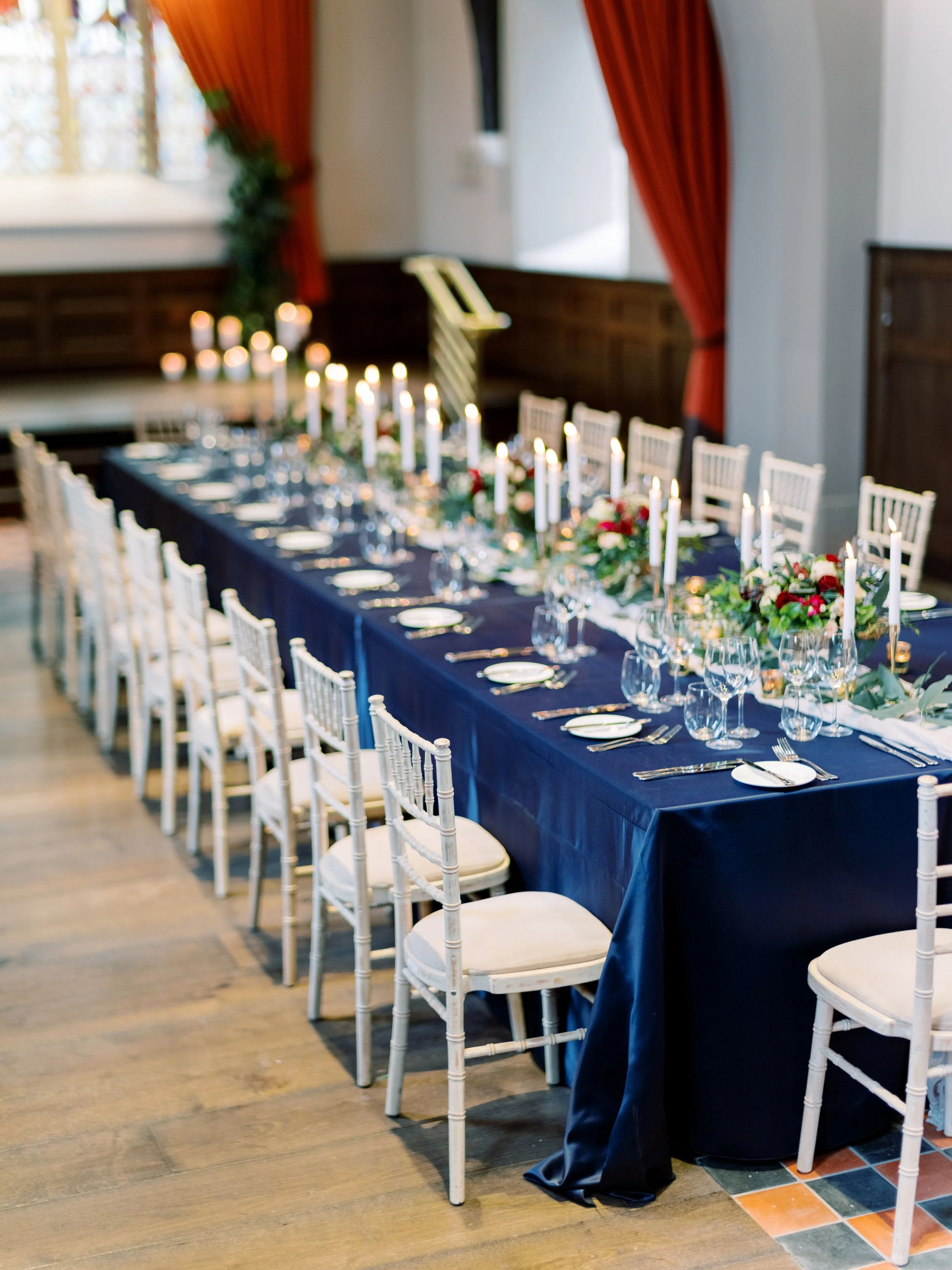 A long banquet table decorated for a formal event with a navy tablecloth, white chairs, floral arrangements, and white candles in gold holders inside a decorated room.