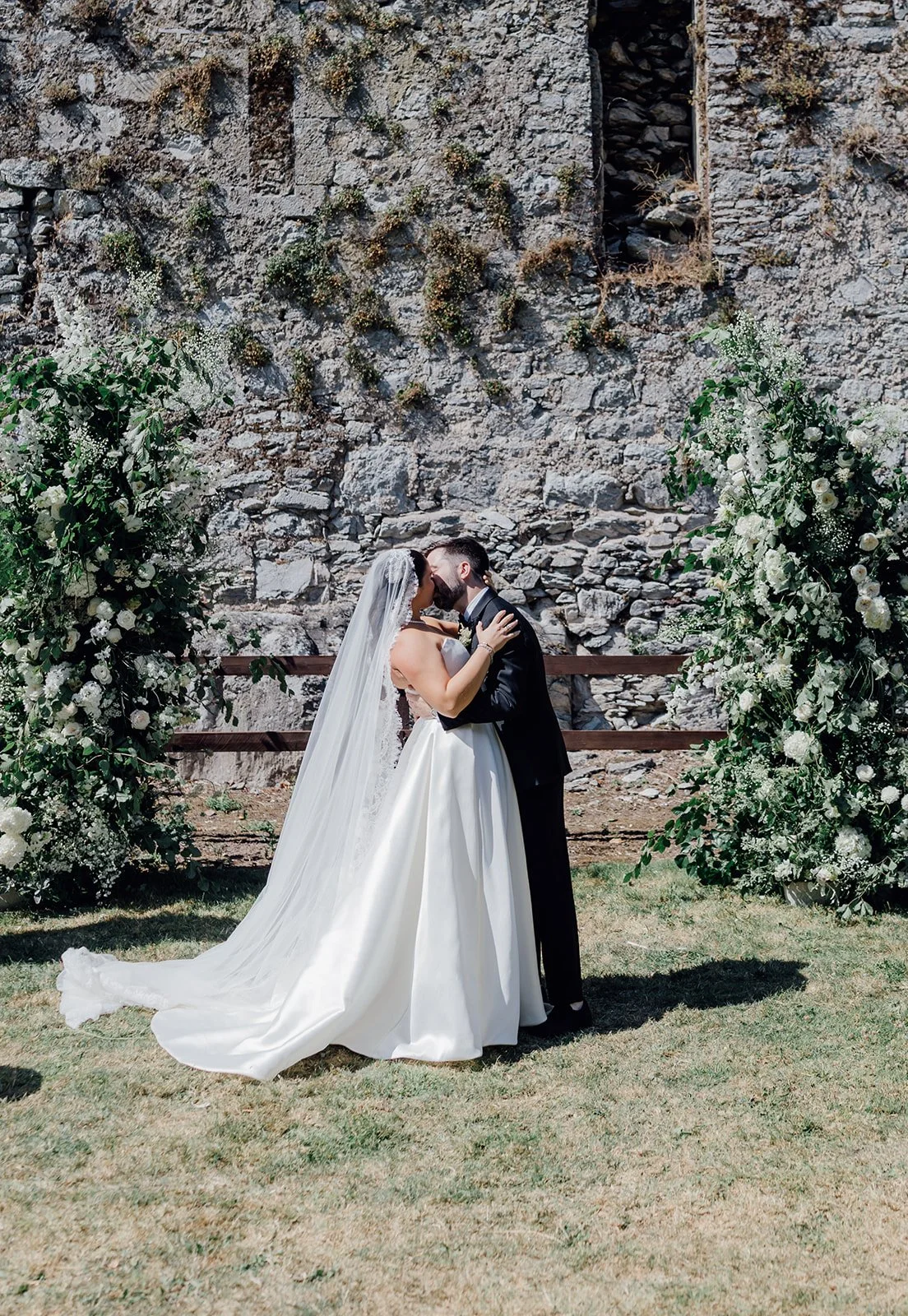 Bride and groom kissing in front of the castle walls and floral arch at Castlemartyr Resort