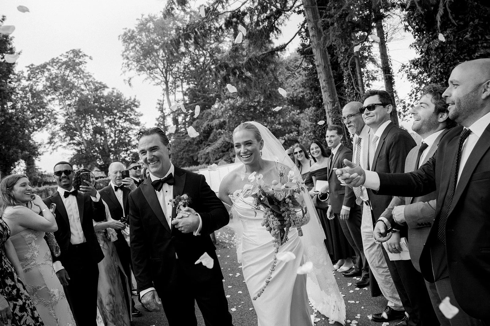 A wedding celebration with a bride in a white gown and veil, holding a bouquet, smiling and walking with a groom in a tuxedo, surrounded by happy guests outdoors during daytime.