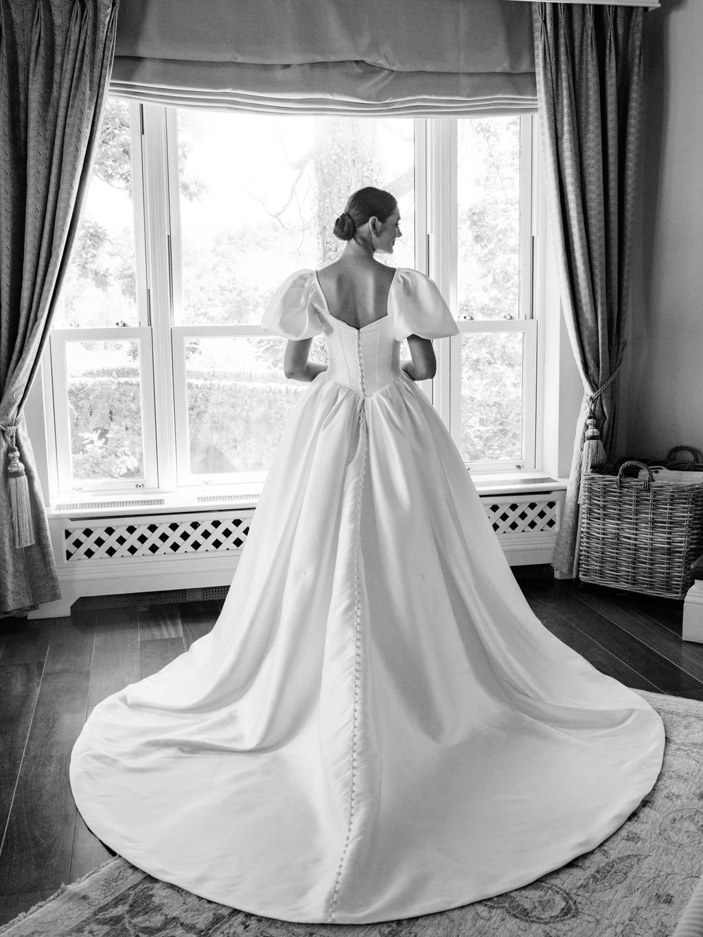 A bride in a wedding gown standing indoors, facing a large window with curtains, in black and white photograph.