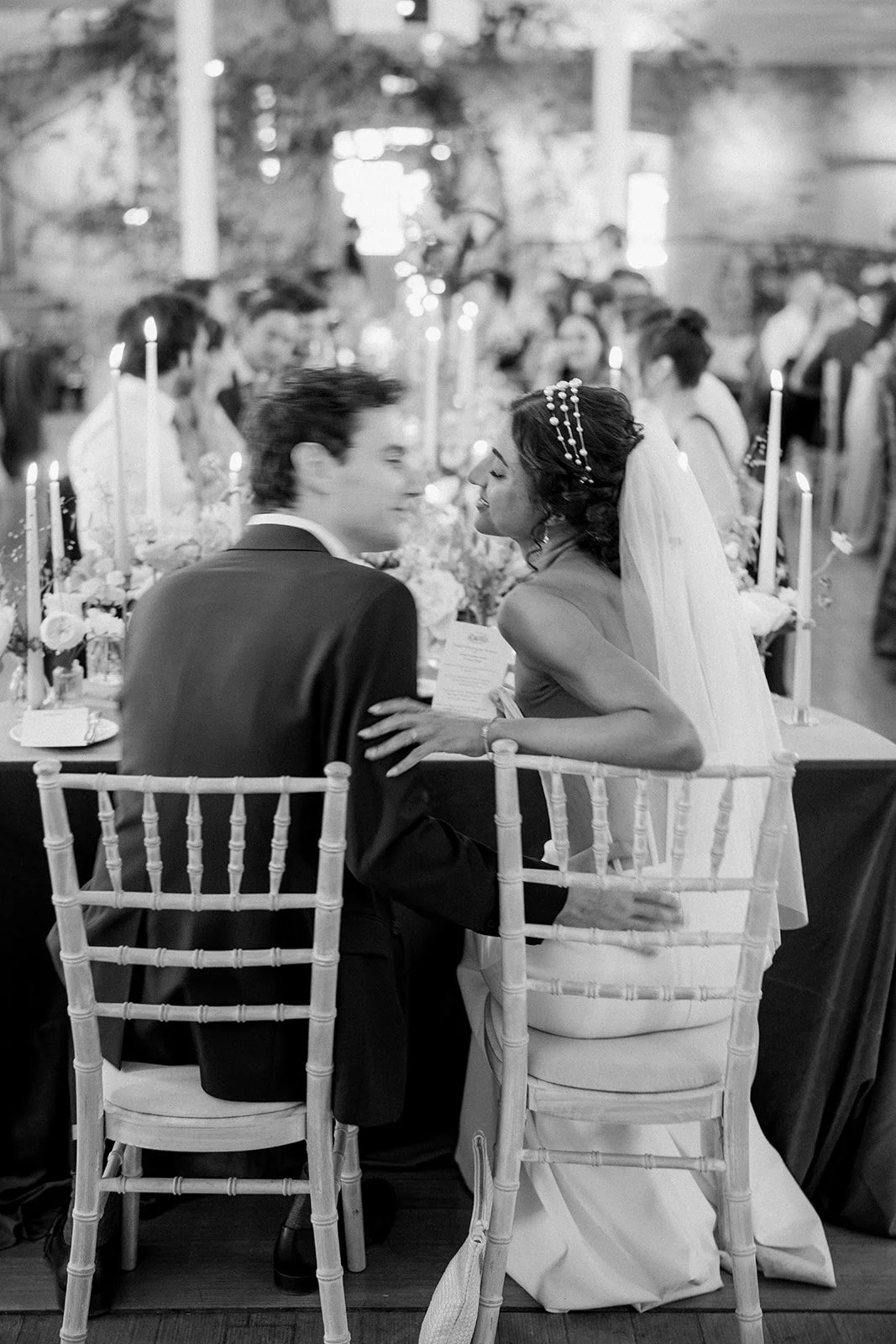 Black and white photo of a bride and groom at their wedding reception, sitting close together at a decorated table with candles, flowers, and guests in the background.