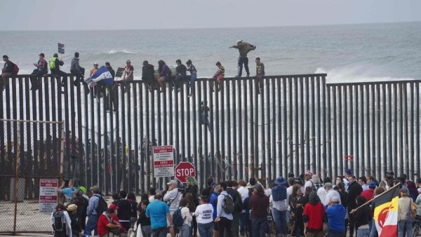 Spectators watch as migrants climb a wall to enter Mexico on their way to the United States.