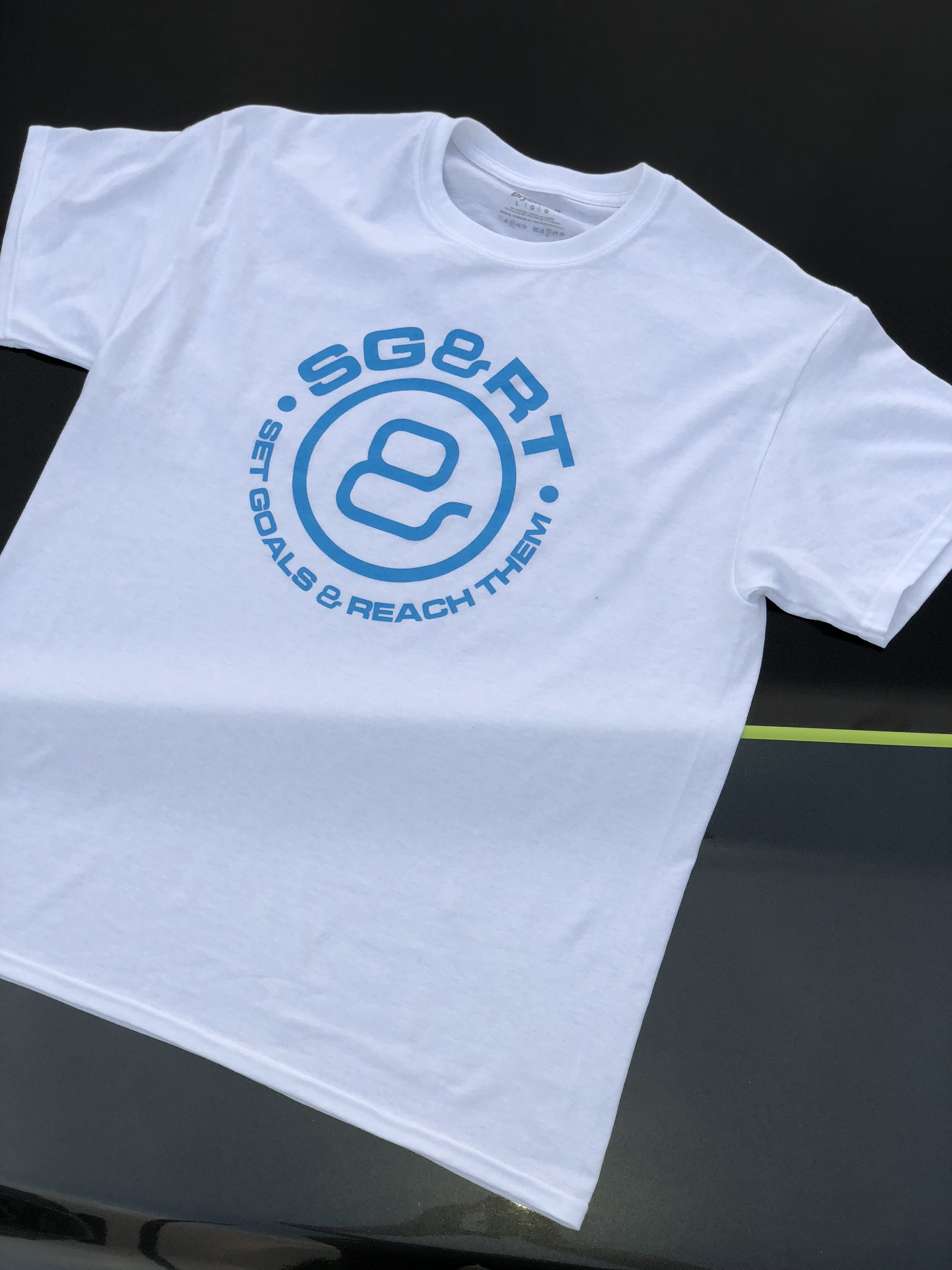 White T-shirt with blue printed logo and text reading "SGRTQ" and "Set Goals & Reach Them" on a dark surface.