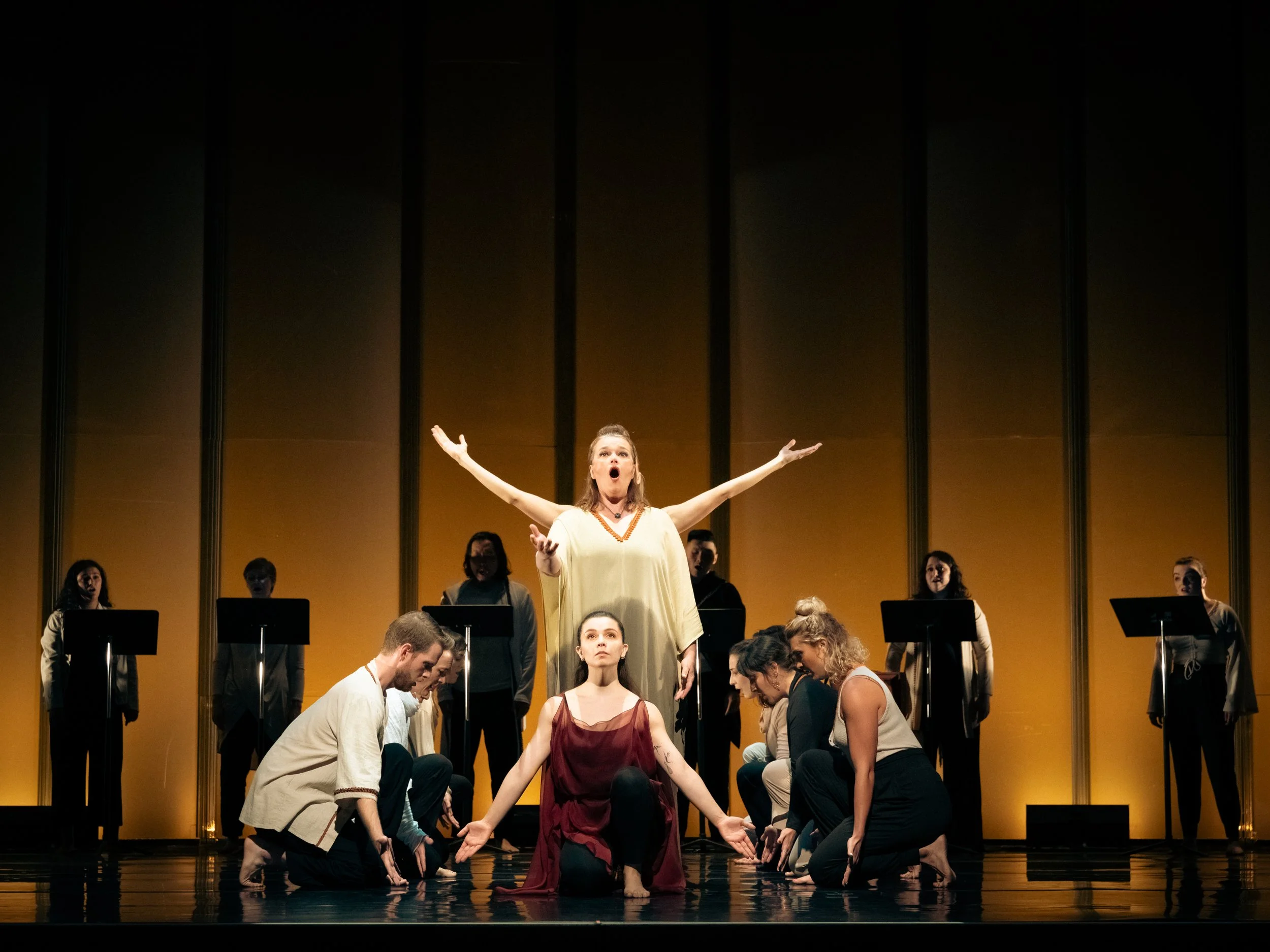 Theater stage with performers, a woman in a beige dress with arms outstretched at center, surrounded by other actors kneeling or sitting, and a choir standing in the background against a yellow backdrop.