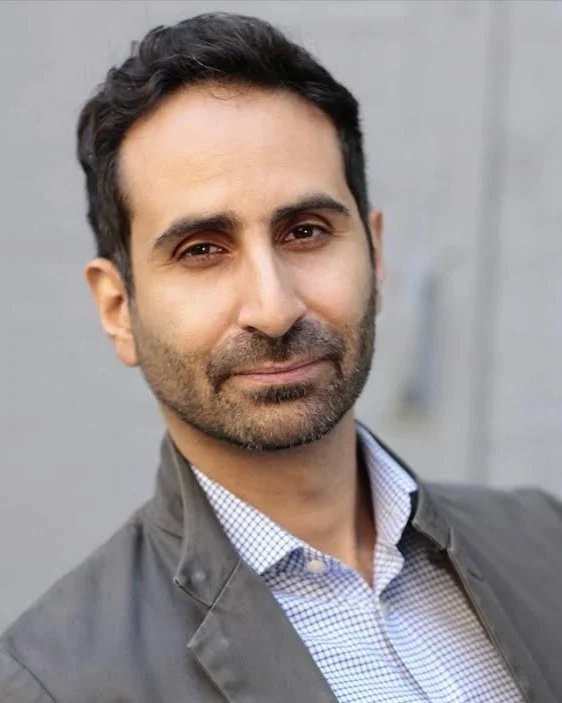 A man with dark hair and a beard, wearing a checkered shirt and a gray jacket, standing outdoors near a gray wall.